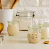 Drei Gläser mit blubberndem Sauerteig, bereit zum Backen. Ein Holzlöffel und eine Schüssel mit Mehl liegen daneben.