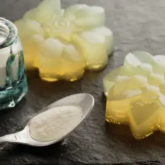 Pulverförmiges Agar Agar Gelatine in einem Glas und auf einem Löffel, daneben leuchtende, blumenförmige Gelees.