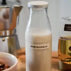 Zutaten für Haselnussmilch selber machen: Haselnüsse, Moka-Kanne, Agavendicksaft und eine Flasche mit selbstgemachter Haselnussmilch.