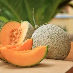 Eine ganze und eine angeschnittene Cantaloupe Melone mit saftigen Scheiben liegen auf einem Holzbrett.