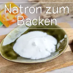 Eine Schale mit weißem Natron zum Backen steht auf einem Holzbrett. Im Hintergrund sind Packungen zu sehen.