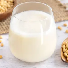 Ein Glas frisch zubereitete Sojamilch, daneben eine Holzschaufel voller Sojabohnen. Sojamilch selber machen ist einfach und gesund.