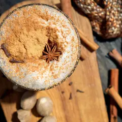 Ein Glas mit Milchschaum, Zimt und Sternanis. Perfekt, um chai selber machen zu lernen. Gewürze wie Zimtstangen und Muskatnüsse liegen daneben.