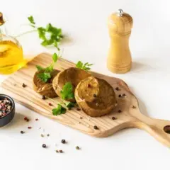 Drei runde seitan steaks liegen auf einem Holzbrett, garniert mit Petersilie. Daneben eine Ölflasche, Pfeffermühle und eine Schale mit Pfefferkörnern.