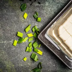 Seidentofu selber machen: Ein Block Seidentofu in einer Schale, daneben geschnittene Frühlingszwiebeln auf dunklem Untergrund.