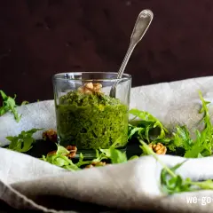 Frisches Rucola Pesto im Glas, garniert mit Walnüssen und Rucolablättern. Ein Löffel steckt darin.
