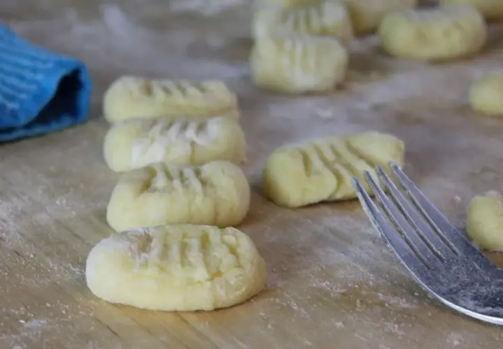 Frisch geformte Gnocchi liegen auf bemehltem Holz. Eine Gabel und ein blauer Lappen deuten darauf hin, dass das Gnocchi selber machen gerade erst begonnen hat.