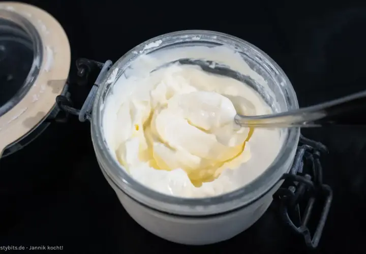 Creme fraiche selber machen ist einfach! Ein Löffel nimmt eine Portion der cremigen Masse aus dem Glas.