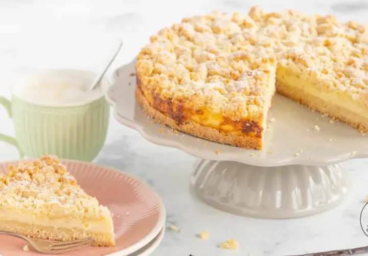 Ein saftiger Streuselkuchen mit Puddingfüllung, bestäubt mit Puderzucker. Daneben eine Tasse Kaffee und ein Stück Kuchen auf einem Teller.