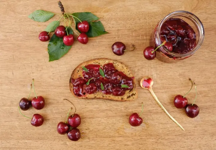 Frisches Brot mit Kirschmarmelade, garniert mit Minze. Ein Glas Kirschmarmelade und frische Kirschen daneben. Perfekt für ein Kirschmarmelade Rezept.