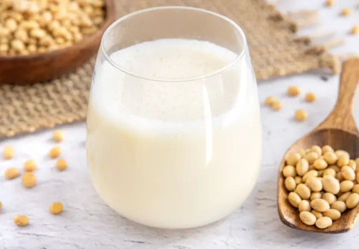 Ein Glas frisch zubereitete Sojamilch, daneben eine Holzschaufel voller Sojabohnen. Sojamilch selber machen ist einfach und gesund.