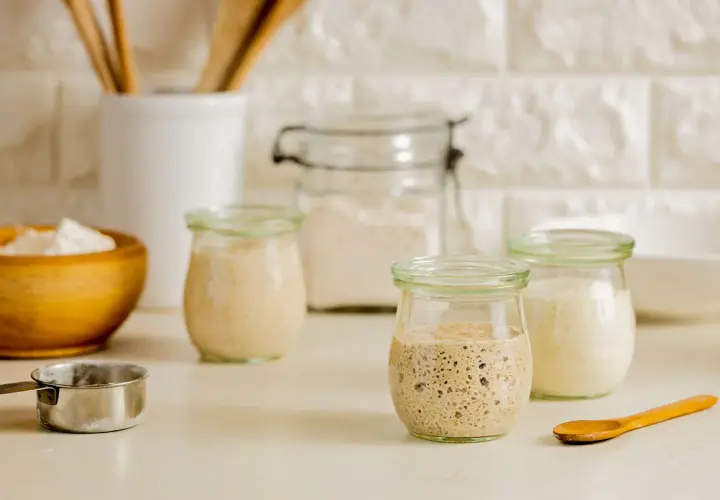 Drei Gläser mit blubberndem Sauerteig, bereit zum Backen. Ein Holzlöffel und eine Schüssel mit Mehl liegen daneben.