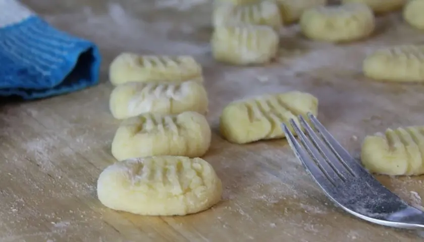 Frisch geformte Gnocchi liegen auf bemehltem Holz. Eine Gabel und ein blauer Lappen deuten darauf hin, dass das Gnocchi selber machen gerade erst begonnen hat.