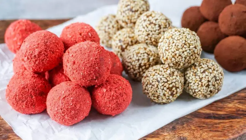 Leckeres Power Balls Rezept: Rote Erdbeer-Bällchen, Sesam-Bällchen und schokoladige Trüffel auf einem Holzbrett.