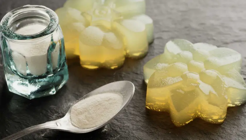 Pulverförmiges Agar Agar Gelatine in einem Glas und auf einem Löffel, daneben leuchtende, blumenförmige Gelees.