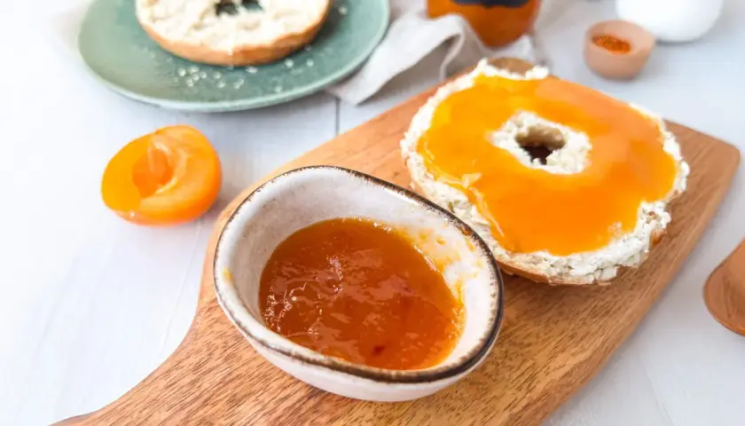 Ein Bagel mit Frischkäse und leckerer Aprikosenmarmelade, die Lust auf das Aprikosenmarmelade Rezept macht. Daneben eine halbe Aprikose.