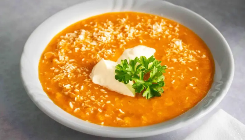 Ein cremiger, orangefarbener rote linsen eintopf, bestreut mit Kokosraspeln und garniert mit einem Klecks Sauerrahm und frischer Petersilie.