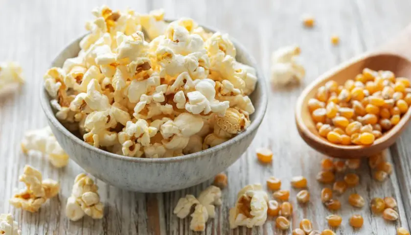 Schüssel mit frisch gemachtem Popcorn, daneben Maiskörner in einem Holzlöffel. So einfach ist Popcorn selber machen!