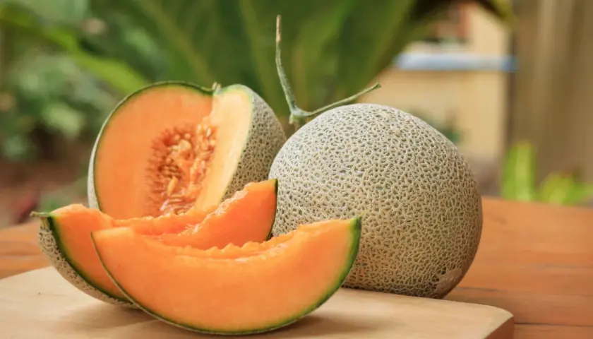 Eine ganze und eine angeschnittene Cantaloupe Melone mit saftigen Scheiben liegen auf einem Holzbrett.