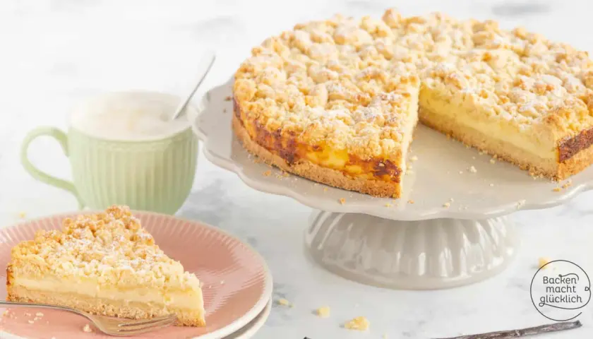 Ein saftiger Streuselkuchen mit Puddingfüllung, bestäubt mit Puderzucker. Daneben eine Tasse Kaffee und ein Stück Kuchen auf einem Teller.