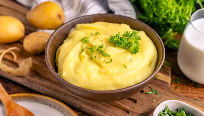Cremiger Kartoffelbrei, garniert mit Petersilie, ist bereit für ein köstliches Kartoffelbrei Rezept. Frische Kartoffeln, Milch und Kräuter liegen bereit.