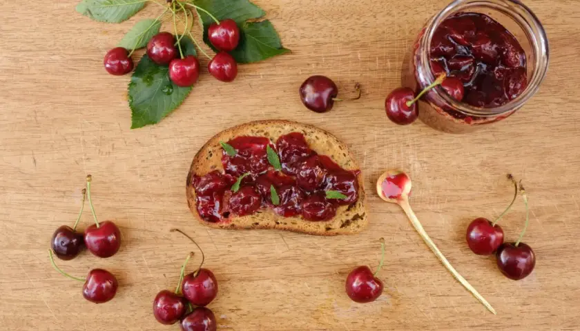 Frisches Brot mit Kirschmarmelade, garniert mit Minze. Ein Glas Kirschmarmelade und frische Kirschen daneben. Perfekt für ein Kirschmarmelade Rezept.