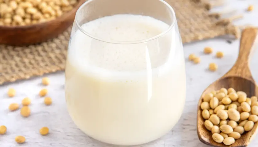 Ein Glas frisch zubereitete Sojamilch, daneben eine Holzschaufel voller Sojabohnen. Sojamilch selber machen ist einfach und gesund.