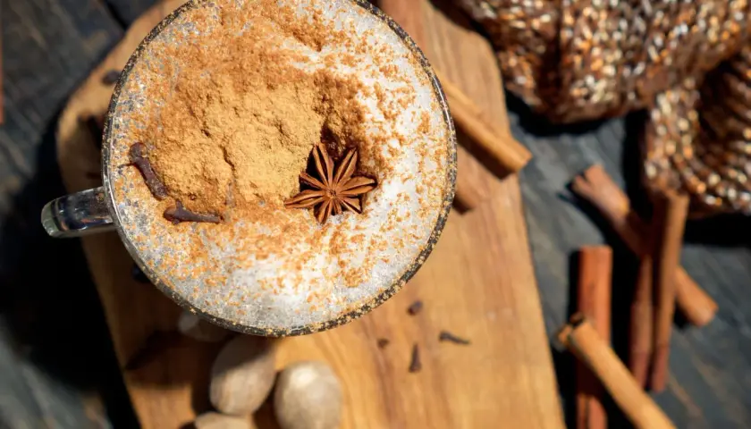 Ein Glas mit Milchschaum, Zimt und Sternanis. Perfekt, um chai selber machen zu lernen. Gewürze wie Zimtstangen und Muskatnüsse liegen daneben.