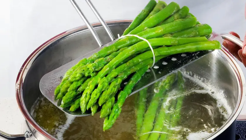 Ein Bund grüner Spargel wird mit einem Abtropfsieb aus kochendem Wasser gehoben. So bereitet man Spargel zu: blanchieren für perfekten Biss.