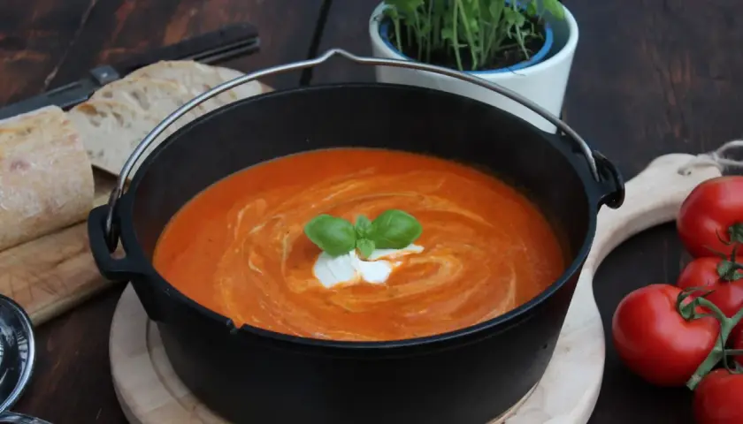 Ein köstliches Tomatensuppe Rezept mit frischen Tomaten, garniert mit Basilikum und einem Klecks Sahne, serviert in einem Gusseisentopf mit Brot.