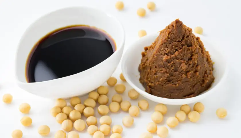 Miso-Paste und Sojasauce in weißen Schalen, umgeben von Sojabohnen. Eine köstliche Alternative zu Hefeflocken für herzhafte Gerichte.