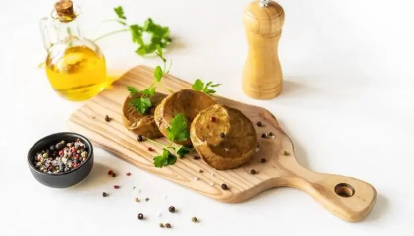 Drei runde seitan steaks liegen auf einem Holzbrett, garniert mit Petersilie. Daneben eine Ölflasche, Pfeffermühle und eine Schale mit Pfefferkörnern.