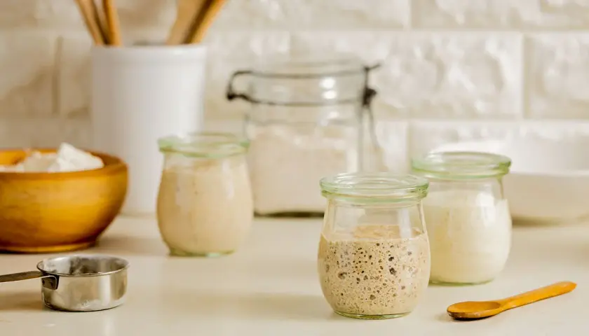 Drei Gläser mit blubberndem Sauerteig, bereit zum Backen. Ein Holzlöffel und eine Schüssel mit Mehl liegen daneben.