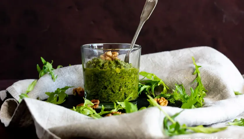 Frisches Rucola Pesto im Glas, garniert mit Walnüssen und Rucolablättern. Ein Löffel steckt darin.