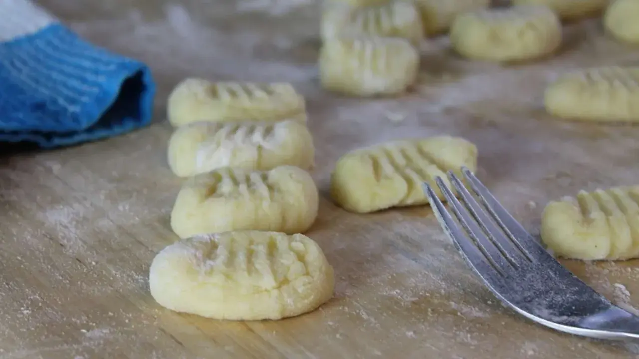 Frisch geformte Gnocchi liegen auf bemehltem Holz. Eine Gabel und ein blauer Lappen deuten darauf hin, dass das Gnocchi selber machen gerade erst begonnen hat.