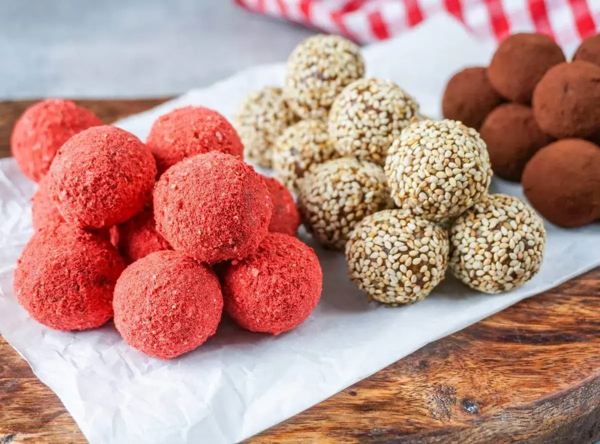 Leckeres Power Balls Rezept: Rote Erdbeer-Bällchen, Sesam-Bällchen und schokoladige Trüffel auf einem Holzbrett.