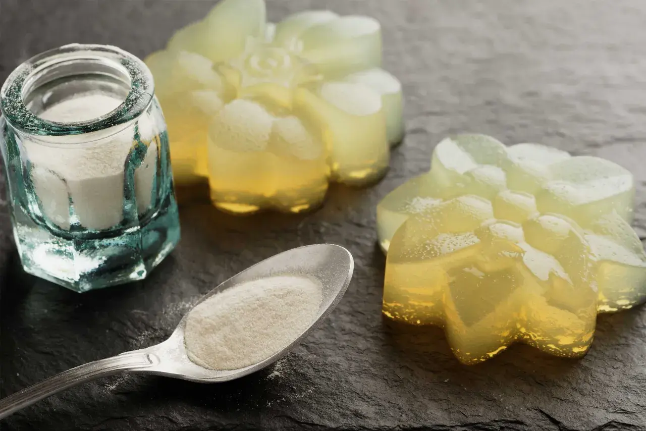 Pulverförmiges Agar Agar Gelatine in einem Glas und auf einem Löffel, daneben leuchtende, blumenförmige Gelees.