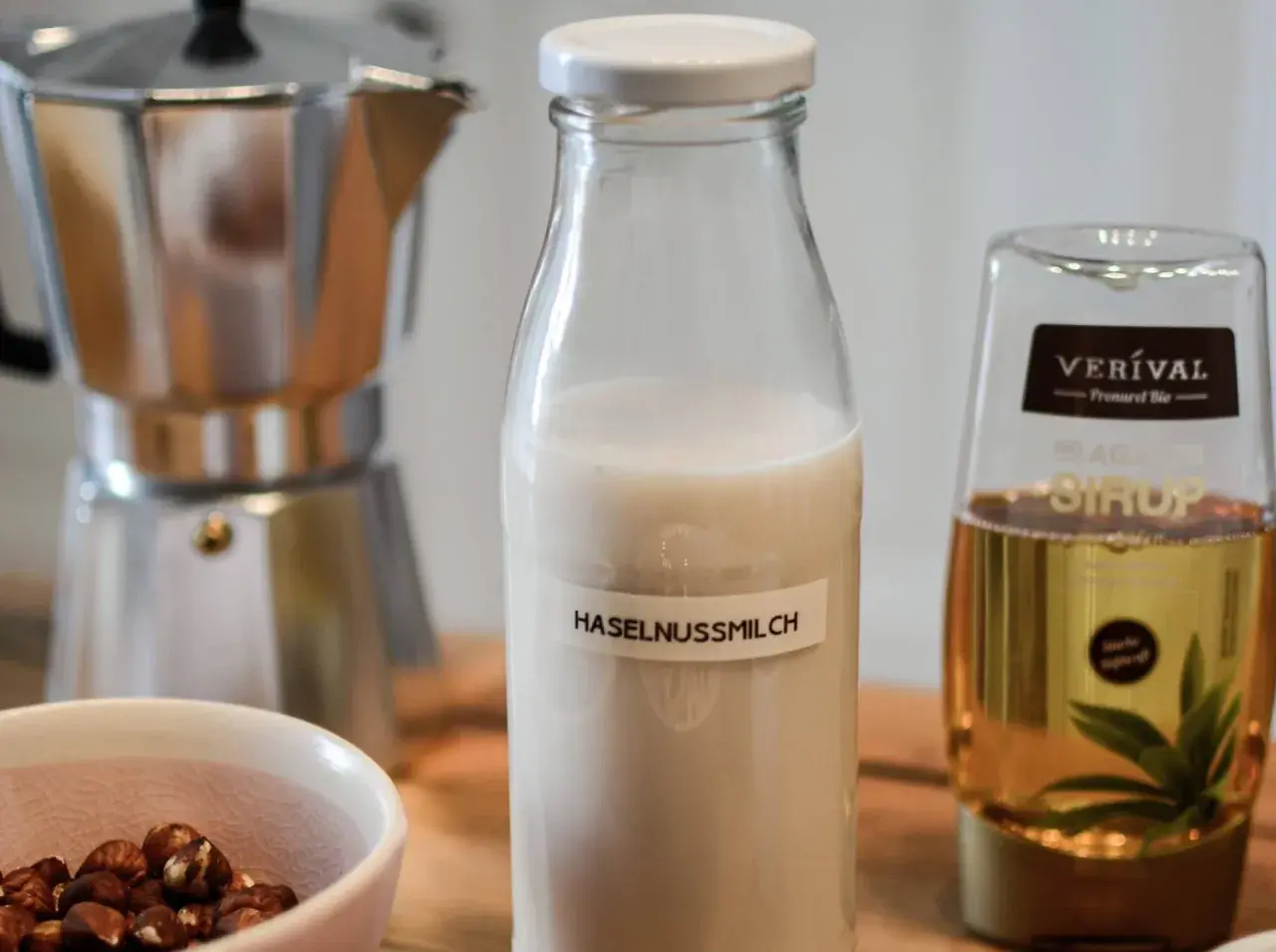 Zutaten für Haselnussmilch selber machen: Haselnüsse, Moka-Kanne, Agavendicksaft und eine Flasche mit selbstgemachter Haselnussmilch.