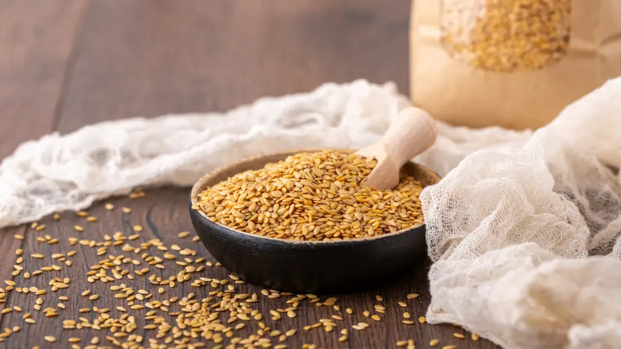 Schale mit goldenen Leinsamen und Holzschaufel, daneben ein Papiersack. Der Unterschied zwischen Leinsamen und Goldleinsamen wird hier visuell dargestellt.