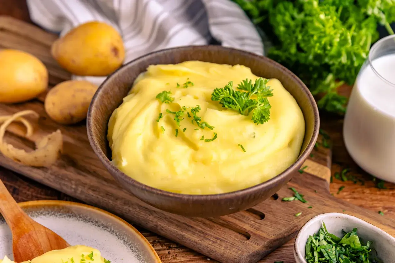 Cremiger Kartoffelbrei, garniert mit Petersilie, ist bereit für ein köstliches Kartoffelbrei Rezept. Frische Kartoffeln, Milch und Kräuter liegen bereit.
