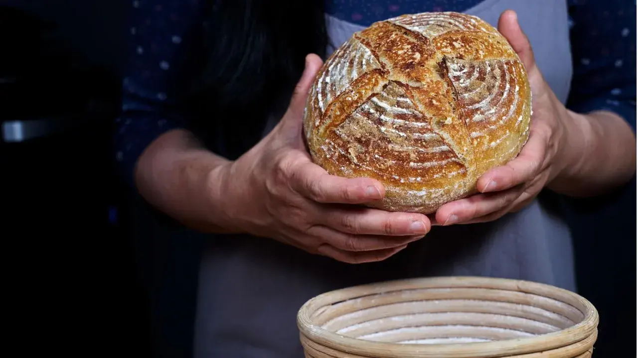 Hände halten ein frisch gebackenes Sauerteigbrot, das gerade aus dem Gärkorb genommen wurde. Der Teig ist perfekt aufgegangen.