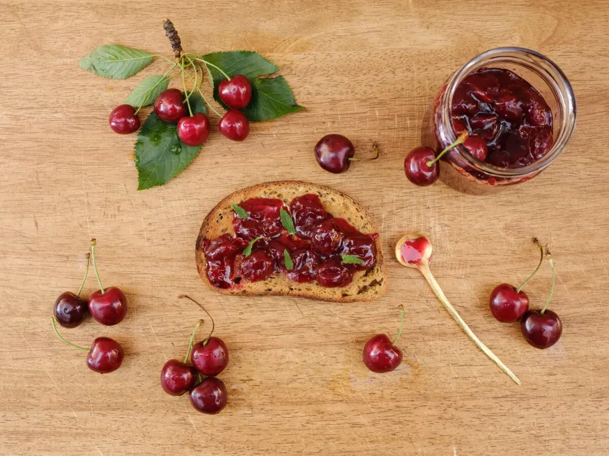 Frisches Brot mit Kirschmarmelade, garniert mit Minze. Ein Glas Kirschmarmelade und frische Kirschen daneben. Perfekt für ein Kirschmarmelade Rezept.