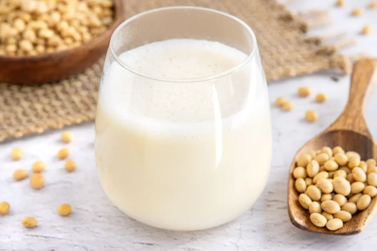 Ein Glas frisch zubereitete Sojamilch, daneben eine Holzschaufel voller Sojabohnen. Sojamilch selber machen ist einfach und gesund.