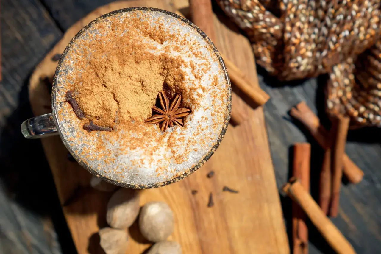 Ein Glas mit Milchschaum, Zimt und Sternanis. Perfekt, um chai selber machen zu lernen. Gewürze wie Zimtstangen und Muskatnüsse liegen daneben.