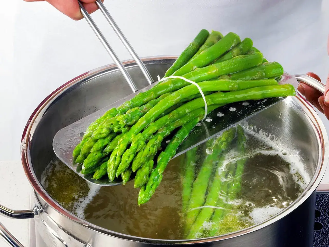 Ein Bund grüner Spargel wird mit einem Abtropfsieb aus kochendem Wasser gehoben. So bereitet man Spargel zu: blanchieren für perfekten Biss.
