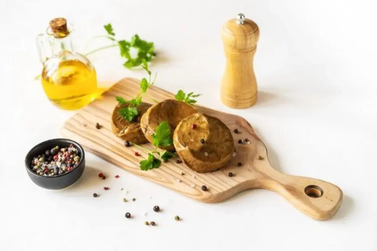 Drei runde seitan steaks liegen auf einem Holzbrett, garniert mit Petersilie. Daneben eine Ölflasche, Pfeffermühle und eine Schale mit Pfefferkörnern.