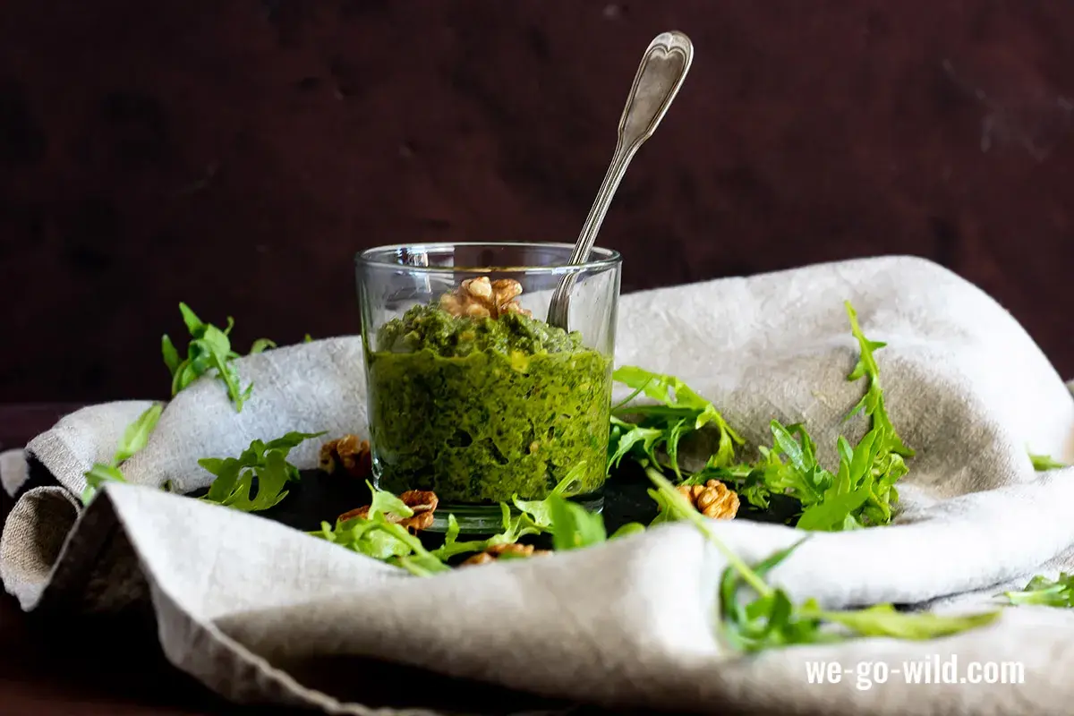 Frisches Rucola Pesto im Glas, garniert mit Walnüssen und Rucolablättern. Ein Löffel steckt darin.