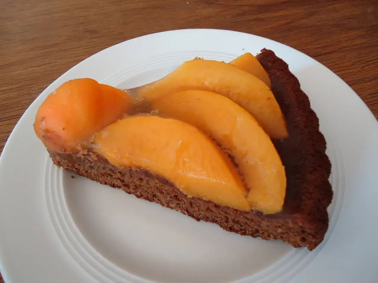 Ein Stück veganer tortenboden mit saftigen Aprikosen, überzogen mit Gelee, auf einem weißen Teller.