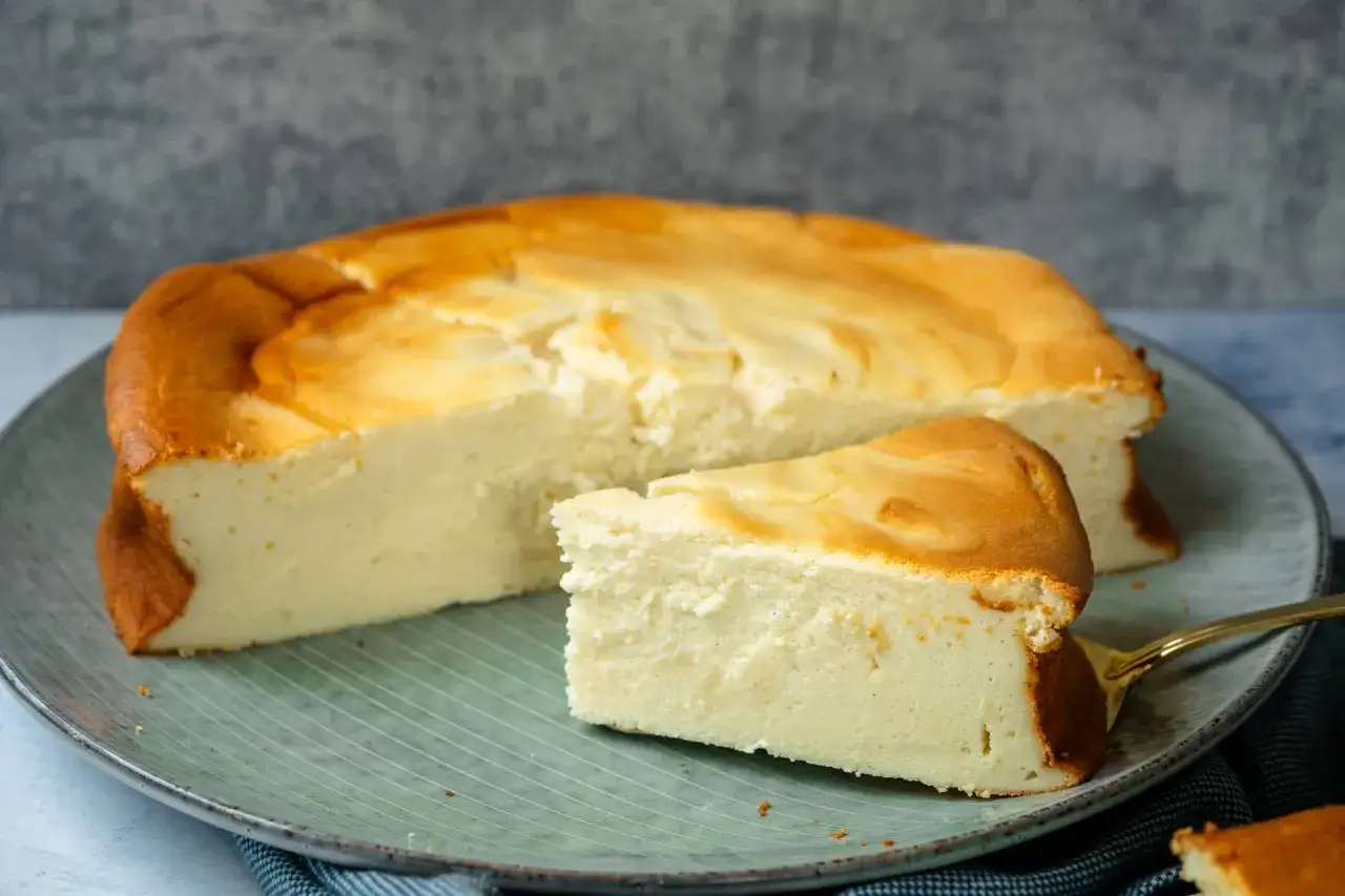 Ein saftiger Quarkkuchen ohne Boden, goldbraun gebacken, liegt auf einem Teller. Ein St&uuml;ck ist bereits angeschnitten.