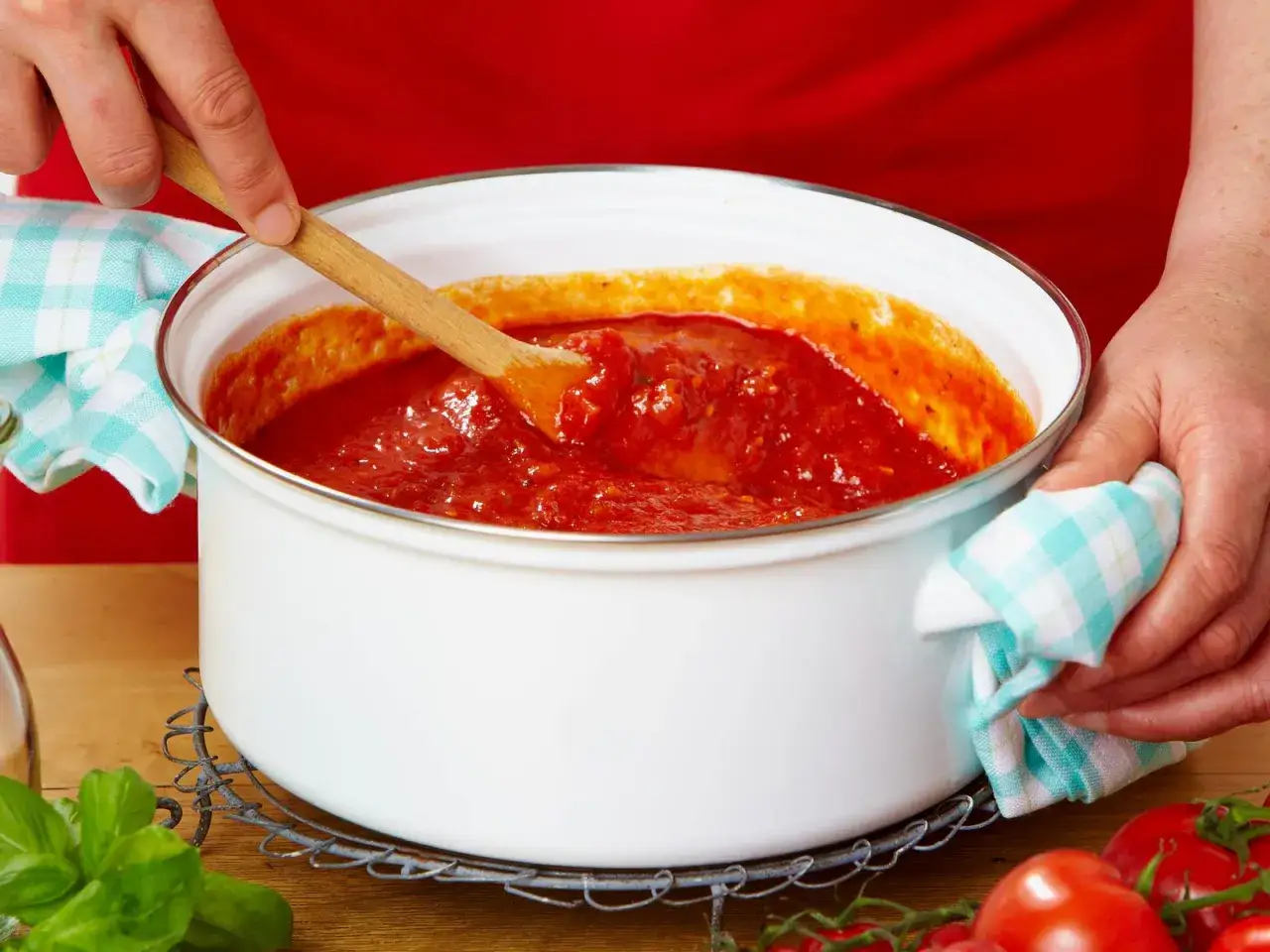 Hände rühren in einem Topf mit köstlicher, selbstgemachter Tomatensauce. Frische Tomaten und Basilikum liegen daneben.
